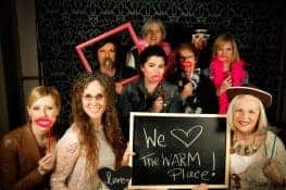 A playful portrait of a group of women wearing silly masks and holding a sign that says "We love The WARM Place!"