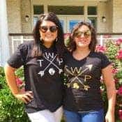 Two women embrace and smile for the camera while modeling black The WARM Place depicting crossed arrows.